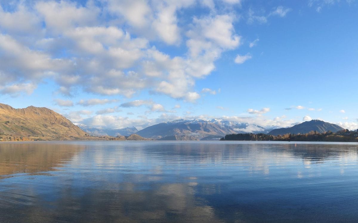 Still water and mountain ranges across Lake Wānaka under a broken-cloud sky